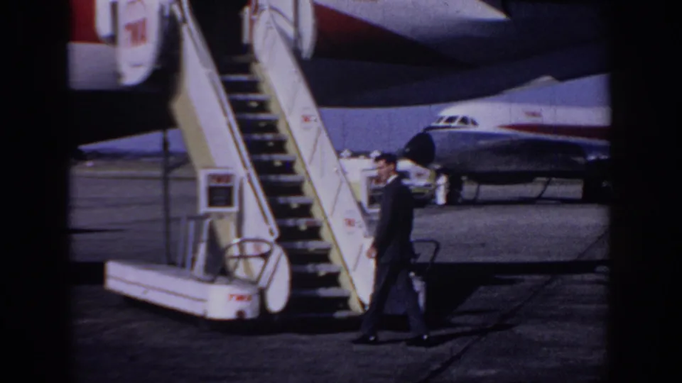 A suited man carries a bag near mobile boarding stairs, with TWA aircraft markings visible behind him on the airport tarmac.