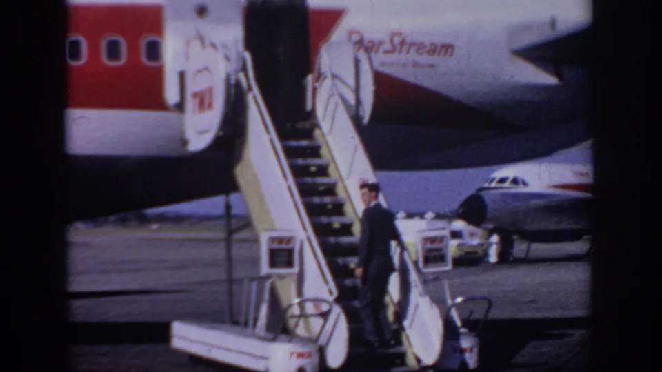 The suited passenger reaches the base of the mobile boarding stairs beside a TWA aircraft, with a yellow service van nearby.