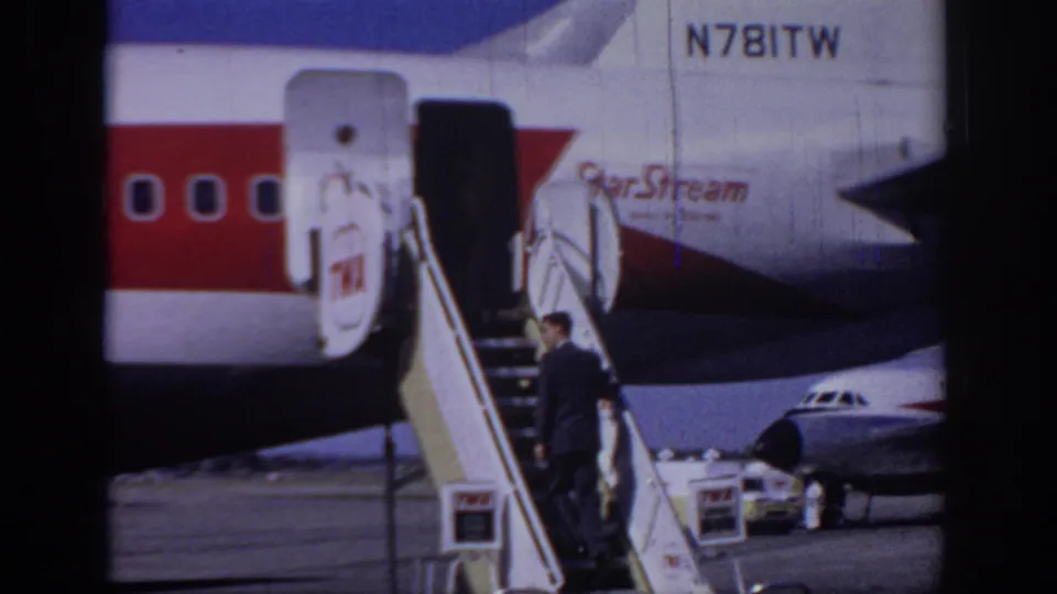 A man in a dark suit is seen beside the boarding stairs of a TWA StarStream plane, with tail registration N781TW visible.