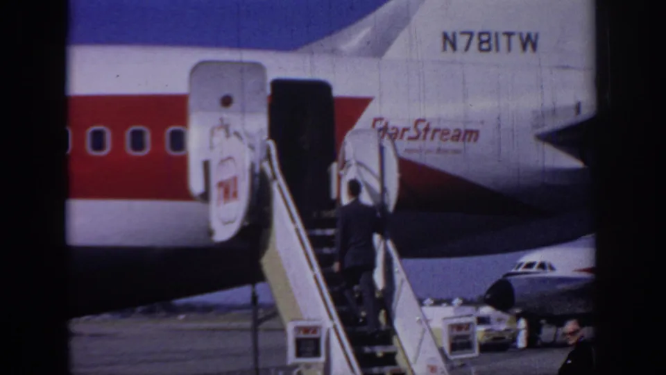 1966 airport frame showing the suited passenger ascending mobile stairs toward the open door of the TWA StarStream aircraft.