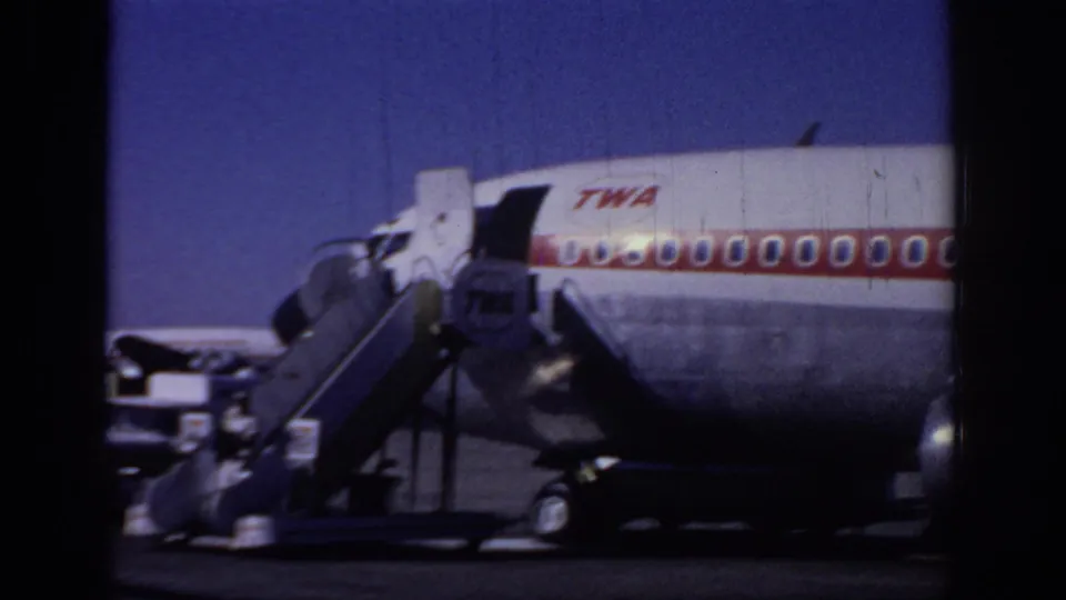 A TWA jet sits on the tarmac with mobile stairs at the forward door and a red-striped fuselage visible in the frame.
