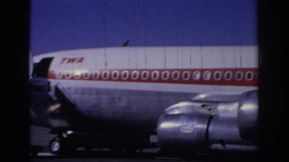 The frame shows the side of a TWA jet with a red-striped fuselage, rows of windows, landing gear area, and a visible engine.