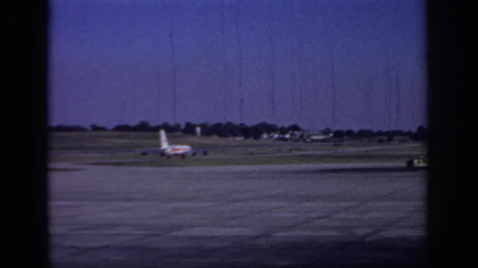 1966 Sacramento home-movie frame showing a distant aircraft on the runway with open tarmac, low horizon structures, and blue sky.