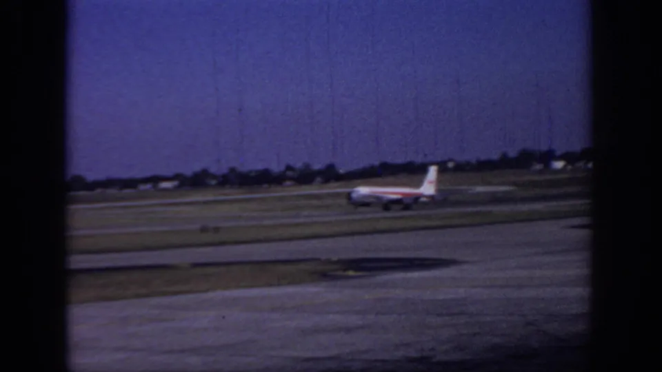 A commercial airliner with red-striped livery moves along the Sacramento runway, with grass fields and low airport structures in the background.