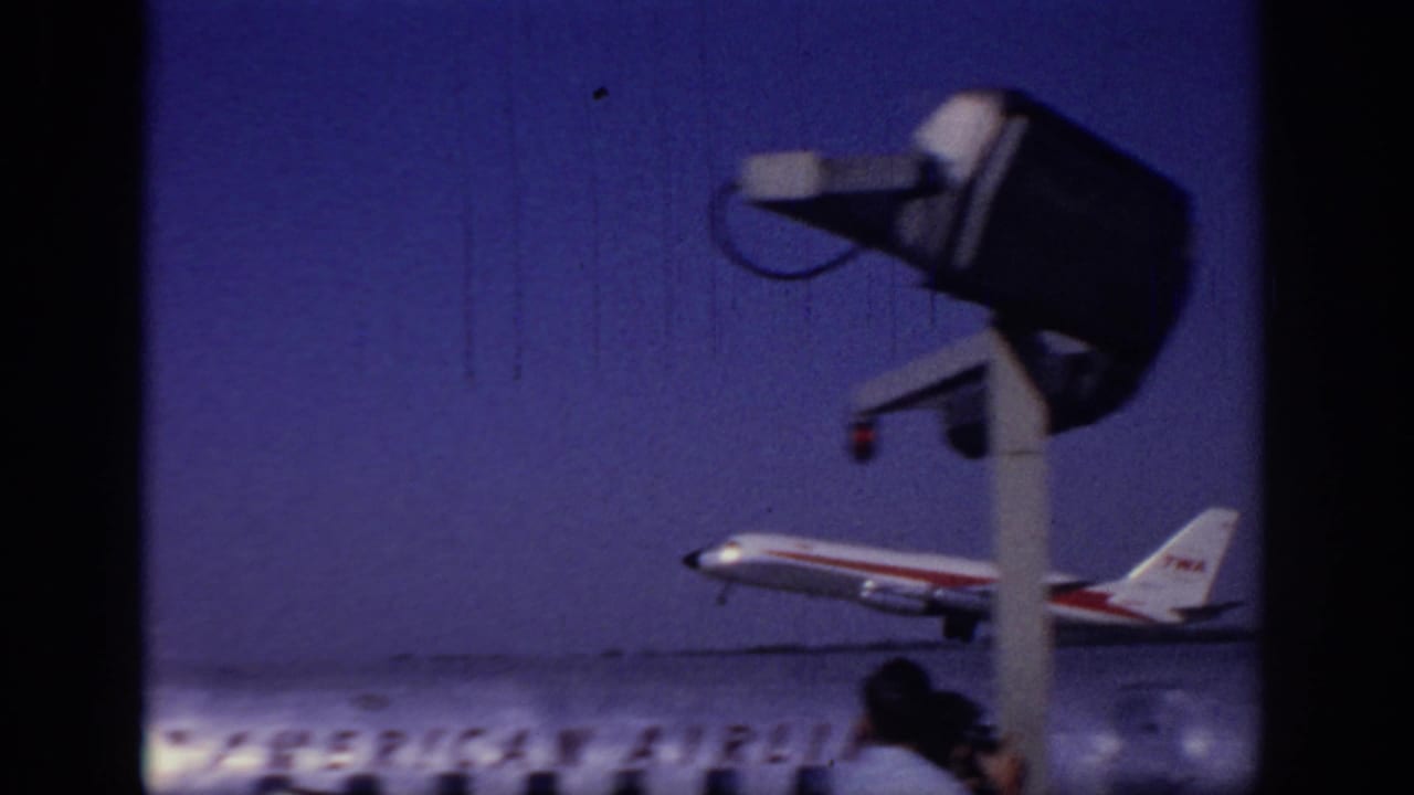 TWA jet lifting from the runway with airport equipment and spectators below.