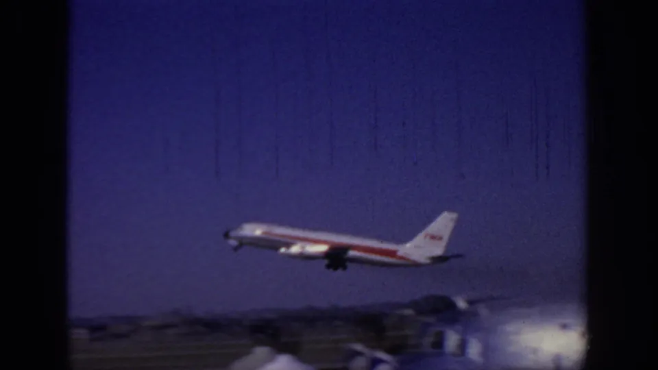 1966 Sacramento home-movie frame showing a TWA airliner climbing above the runway with airport structures and distant low hills below.