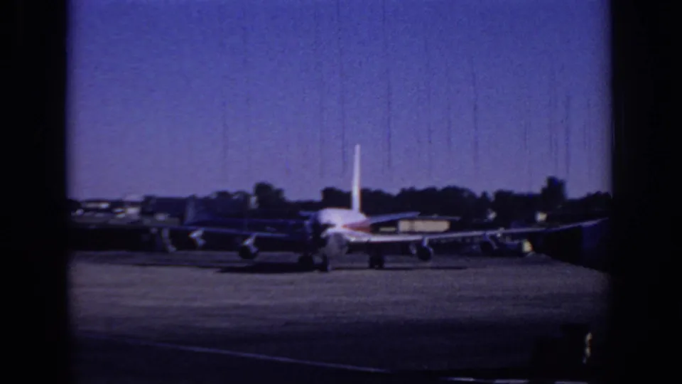 1966 Sacramento home-movie frame showing the rear of a passenger jet on the airport apron, with parked aircraft and a low tree line beyond.