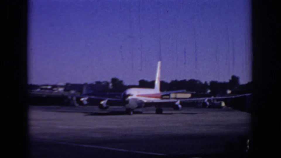 1966 Sacramento airport footage frame with a passenger jet seen from behind on the tarmac, bordered by dark 8mm film edges.