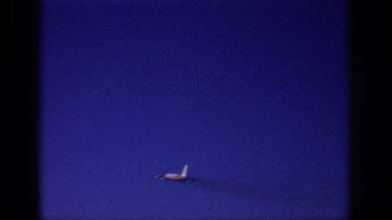 Distant small airplane over Sacramento sky in 1966 home-movie footage, with a faint smoke or vapor trail and visible 8mm grain.