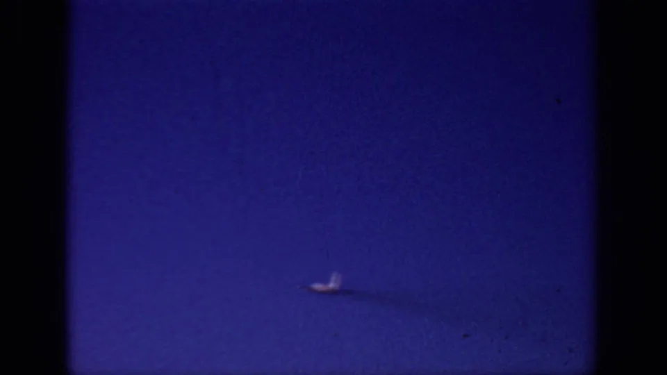A distant airplane sits low in the 1966 Sacramento frame, filmed through grainy 8mm stock against a deep blue sky.