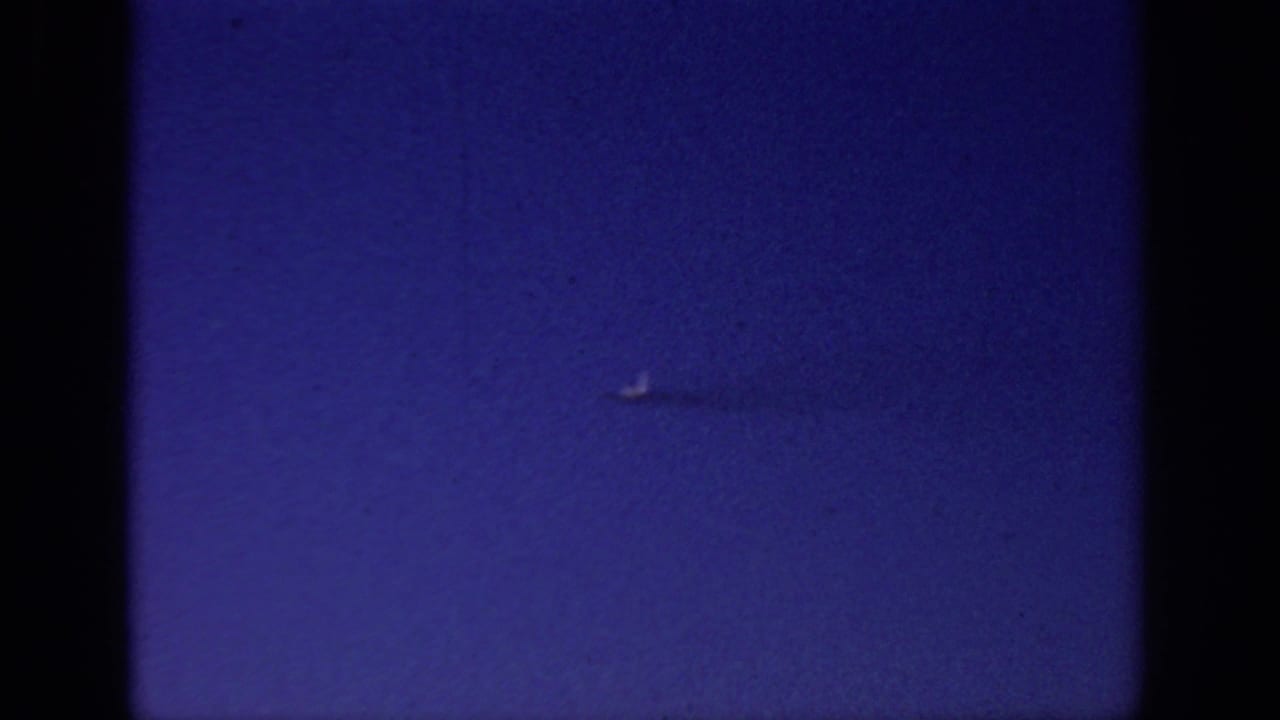 Tiny pale object crossing a deep blue sky with dark 8mm frame edges.