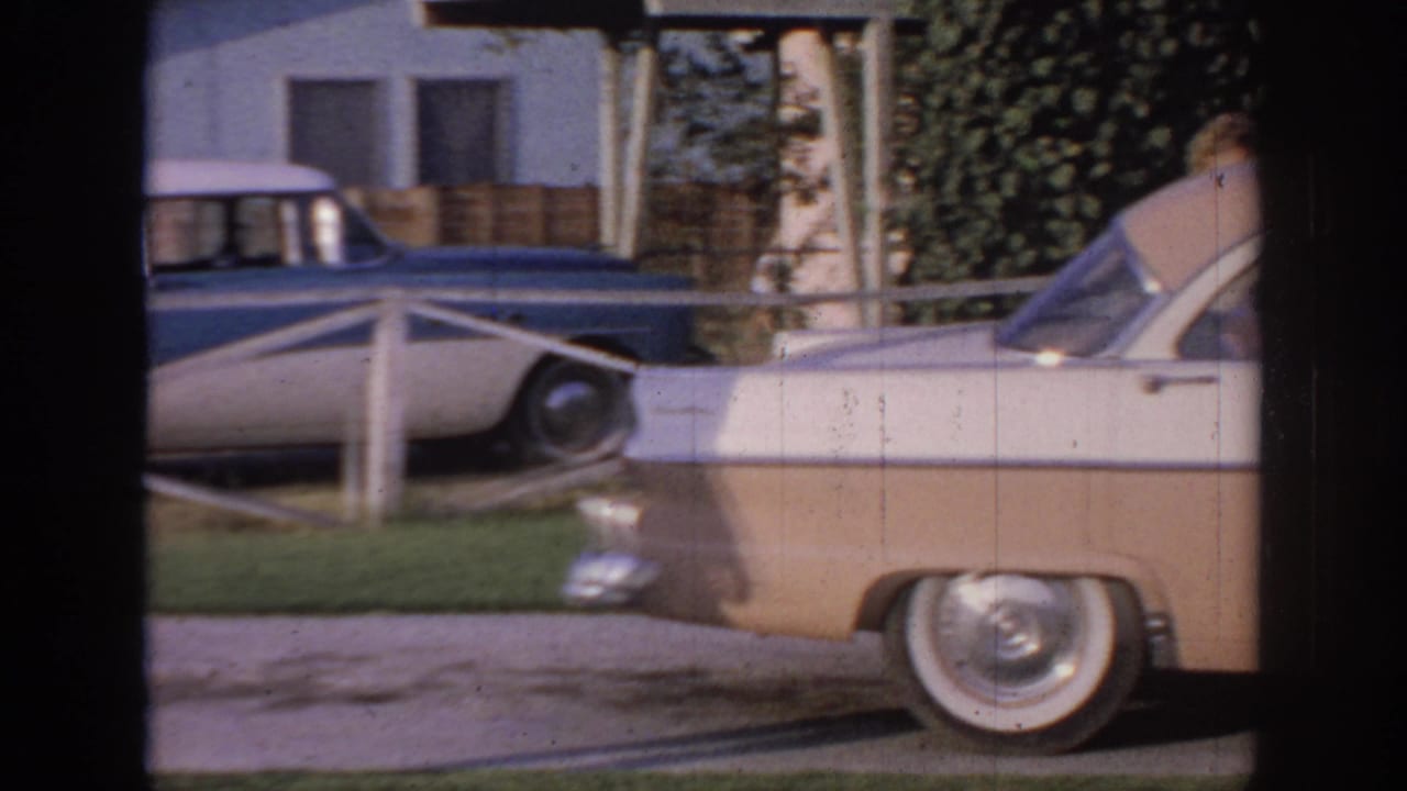 1967 Oklahoma home-movie frame showing a beige-and-white sedan in a suburban driveway with a blue-and-white car near a house behind it.