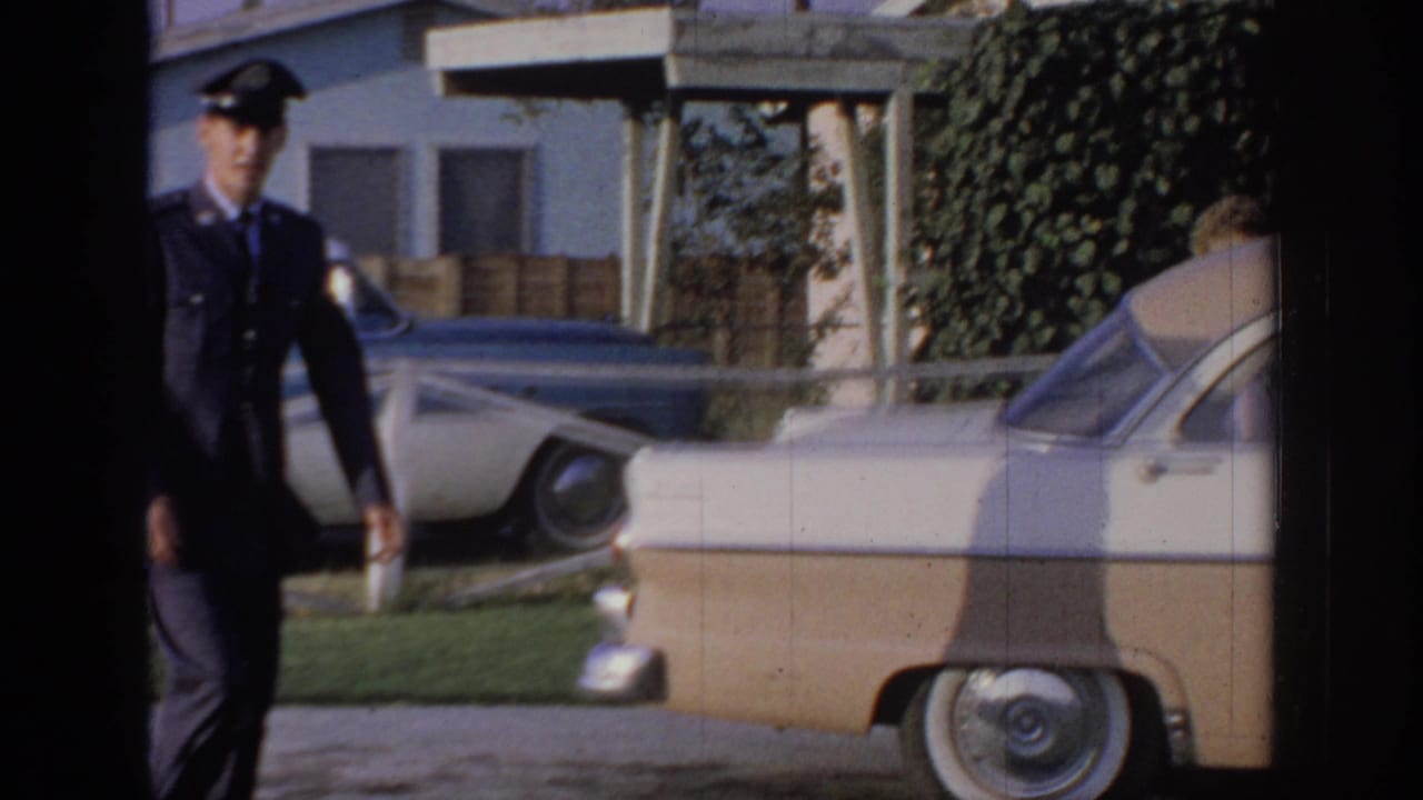1967 Oklahoma home-movie frame with a man in a dark uniform and cap walking toward a parked two-tone sedan outside a house.