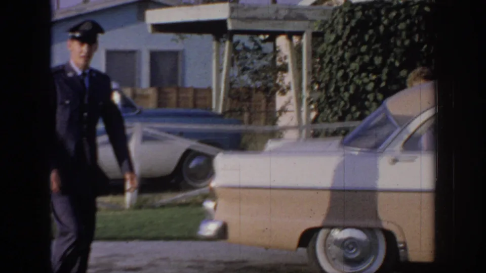 1967 Oklahoma home-movie frame with a man in a dark uniform and cap walking toward a parked two-tone sedan outside a house.
