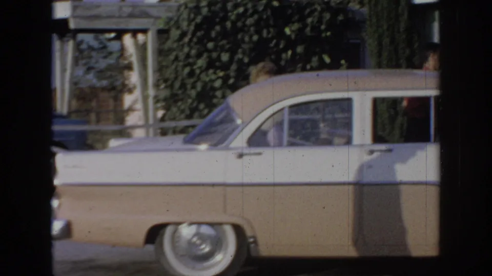 1967 Oklahoma home-movie frame showing the side of a beige-and-white sedan beside a house, with a partly visible person near the car.