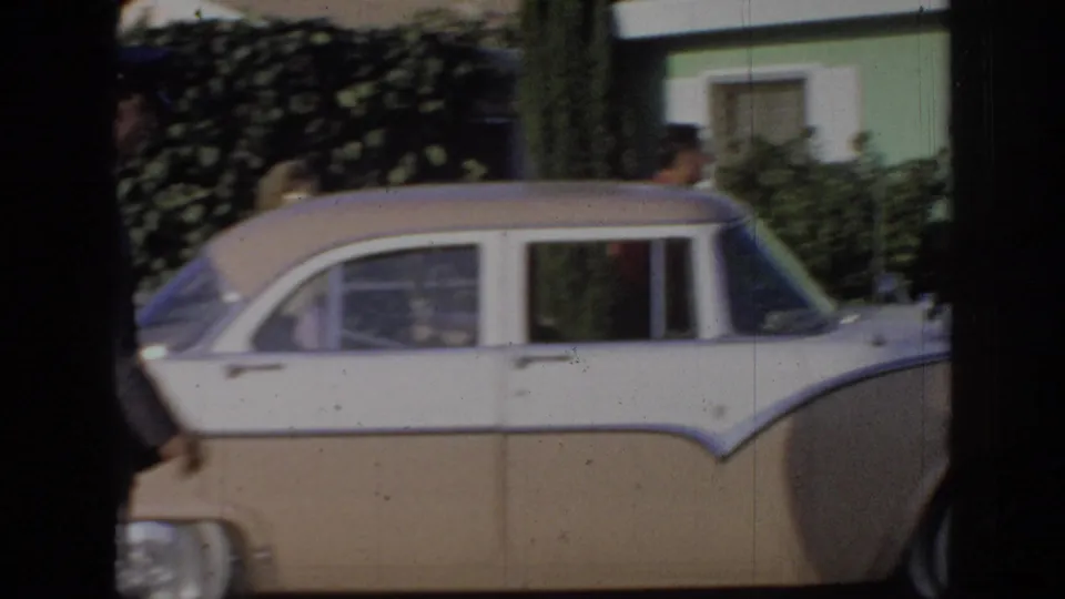 1967 Oklahoma home-movie frame showing a beige-and-white sedan in a residential driveway with shrubbery and a partly visible person behind the car.