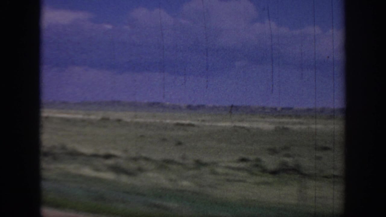 1967 Oklahoma 8mm road footage showing flat green-brown prairie from inside a moving vehicle, with a low horizon, clouded blue-purple sky, and dark window edges.
