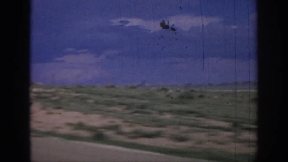 1967 Oklahoma 8mm frame showing low prairie and sandy roadside foreground from a moving car, with a broad blue-purple sky and visible film wear.