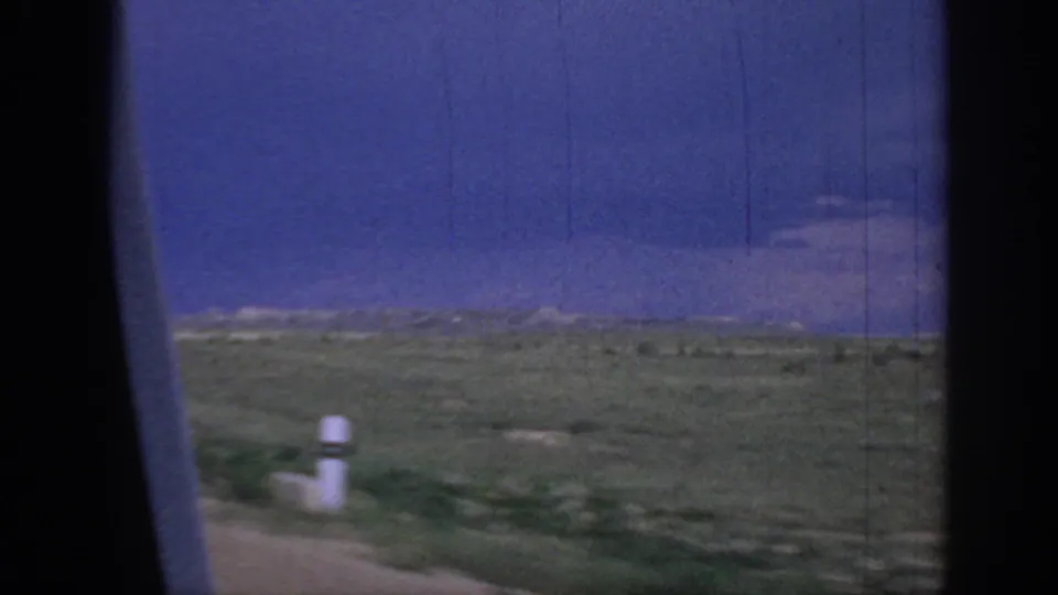 1967 Oklahoma road footage showing flat prairie and a small white roadside post, filmed past a dark car-window edge under a deep blue-purple sky.