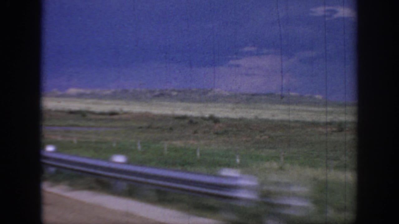 Metal guardrail blurs past open fields under a blue sky.