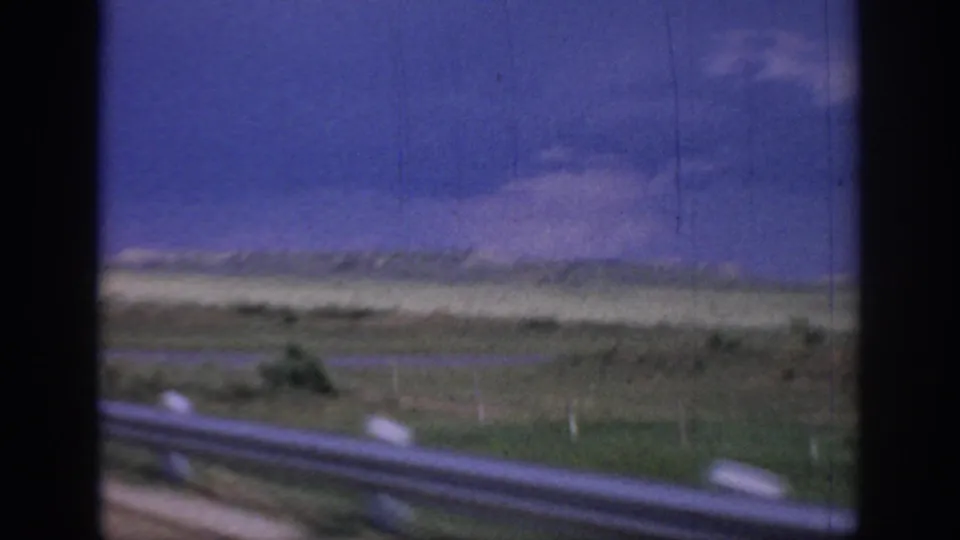 Moving-car view across Oklahoma plains, with a guardrail in the foreground and soft clouds over the distant horizon.