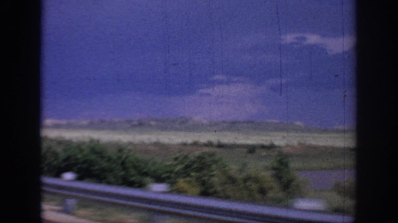 1967 Oklahoma 8mm frame from a moving vehicle, showing a guard rail, roadside brush, flat grassland, distant low hills, and a blue sky with clouds.
