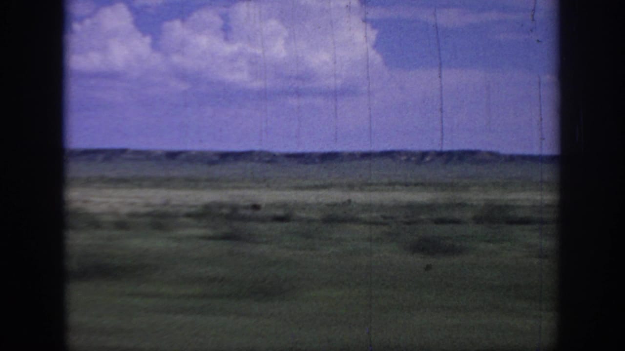 1967 Oklahoma 8mm frame showing open grassland and distant low ridges under a blue sky, viewed through dark vertical frame or window edges.