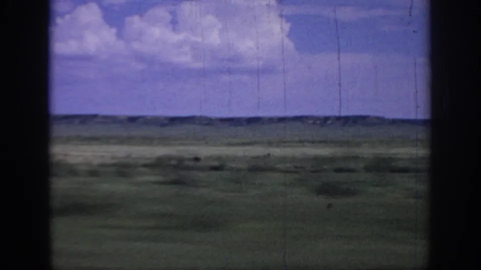 1967 Oklahoma 8mm frame showing open grassland and distant low ridges under a blue sky, viewed through dark vertical frame or window edges.