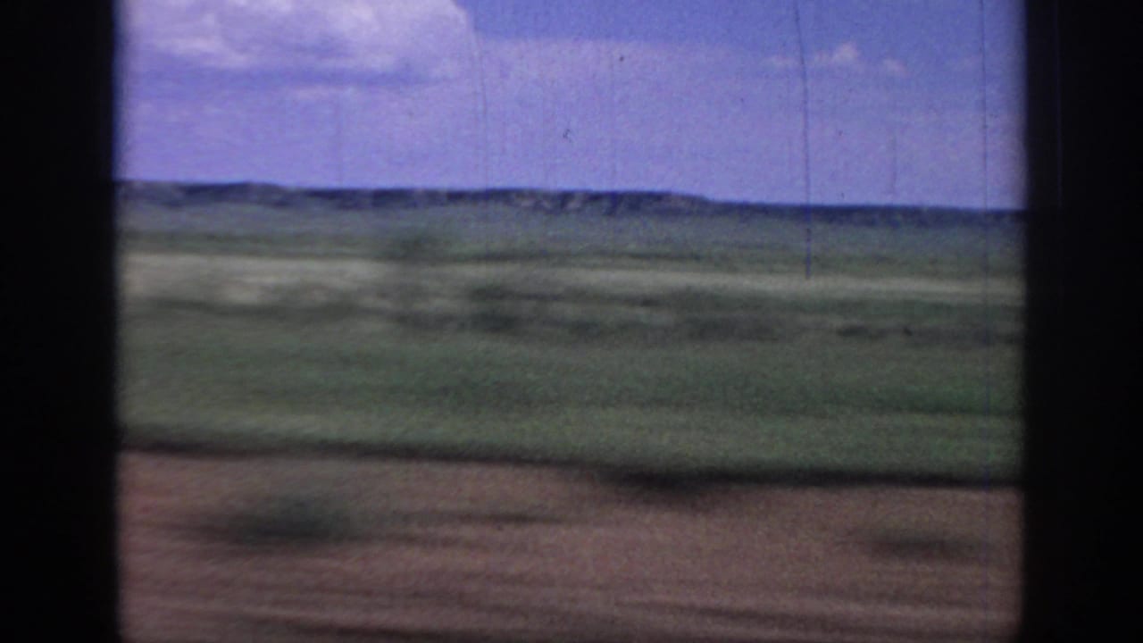 1967 Oklahoma 8mm frame with motion-blurred foreground, green plains, distant hills, blue sky, and visible film scratches from a fast vehicle view.