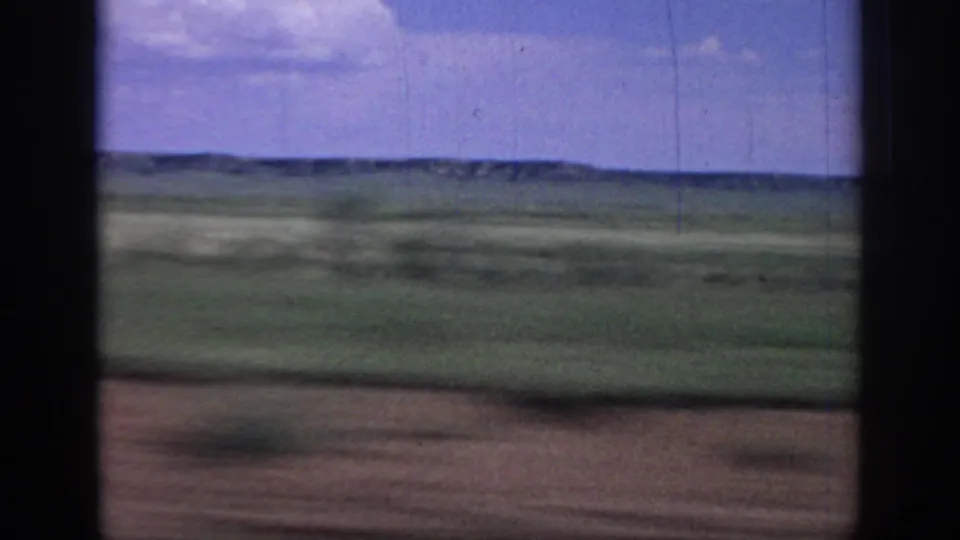 1967 Oklahoma 8mm frame with motion-blurred foreground, green plains, distant hills, blue sky, and visible film scratches from a fast vehicle view.