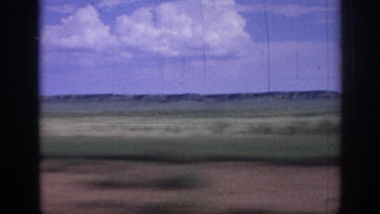 1967 Oklahoma 8mm frame looking across flat grassland toward low distant hills, with white clouds overhead and dark film or window edges at the sides.