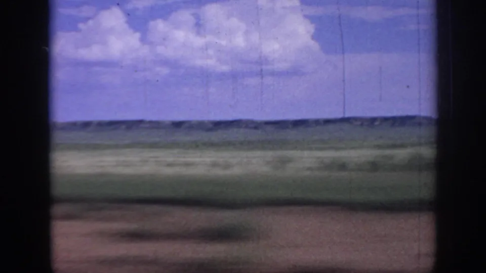 1967 Oklahoma 8mm frame looking across flat grassland toward low distant hills, with white clouds overhead and dark film or window edges at the sides.
