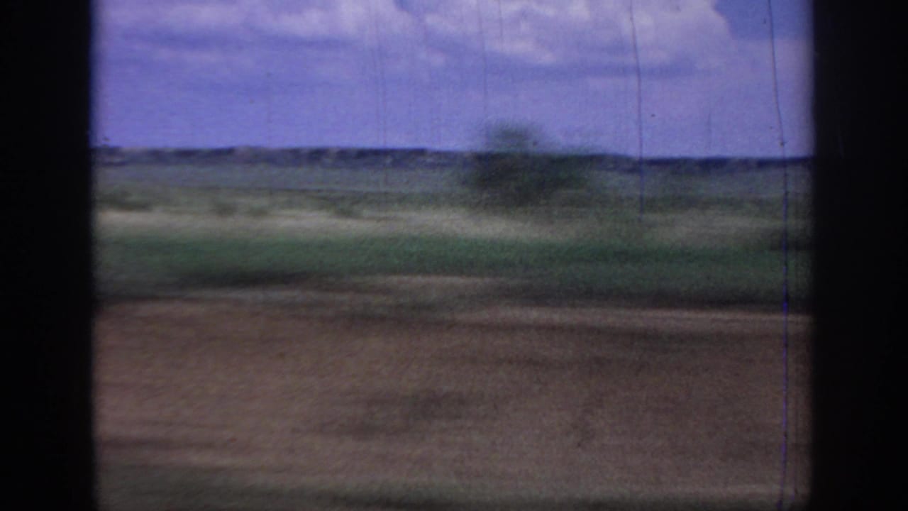 Blurred green and brown fields pass beneath a cloudy blue sky through a window opening.
