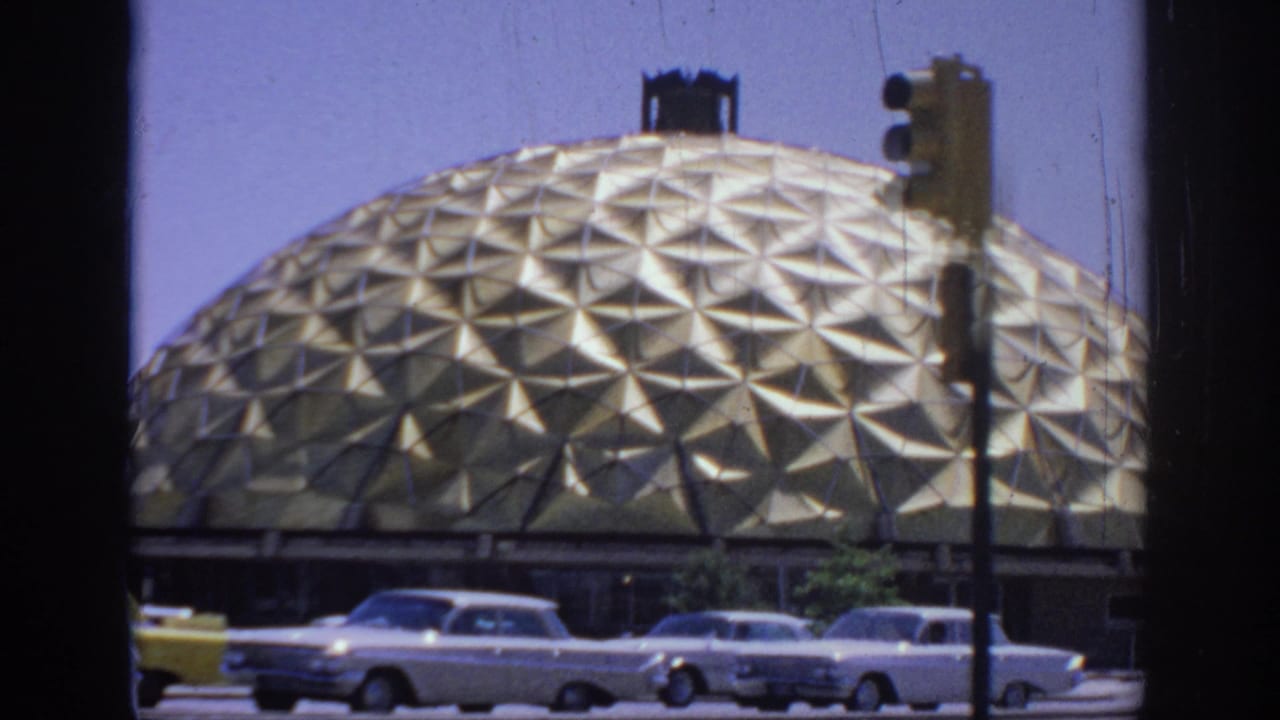 1967 Oklahoma home-movie frame showing a gold triangular-patterned dome beyond a street with sedans passing in front.