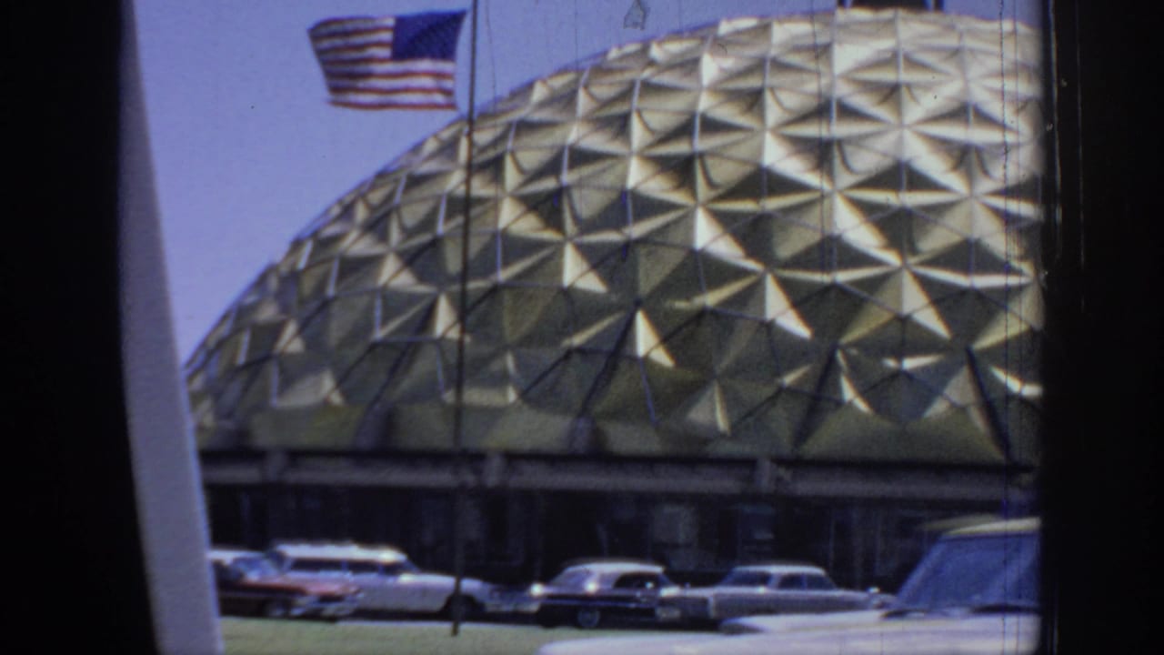 1967 Oklahoma home-movie frame showing a gold triangular-pattern dome, American flag, and classic cars viewed from a vehicle.