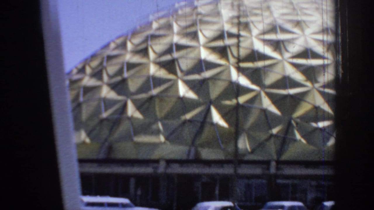 Close vehicle-window view of a gold geodesic dome with parked mid-century cars partly visible below.
