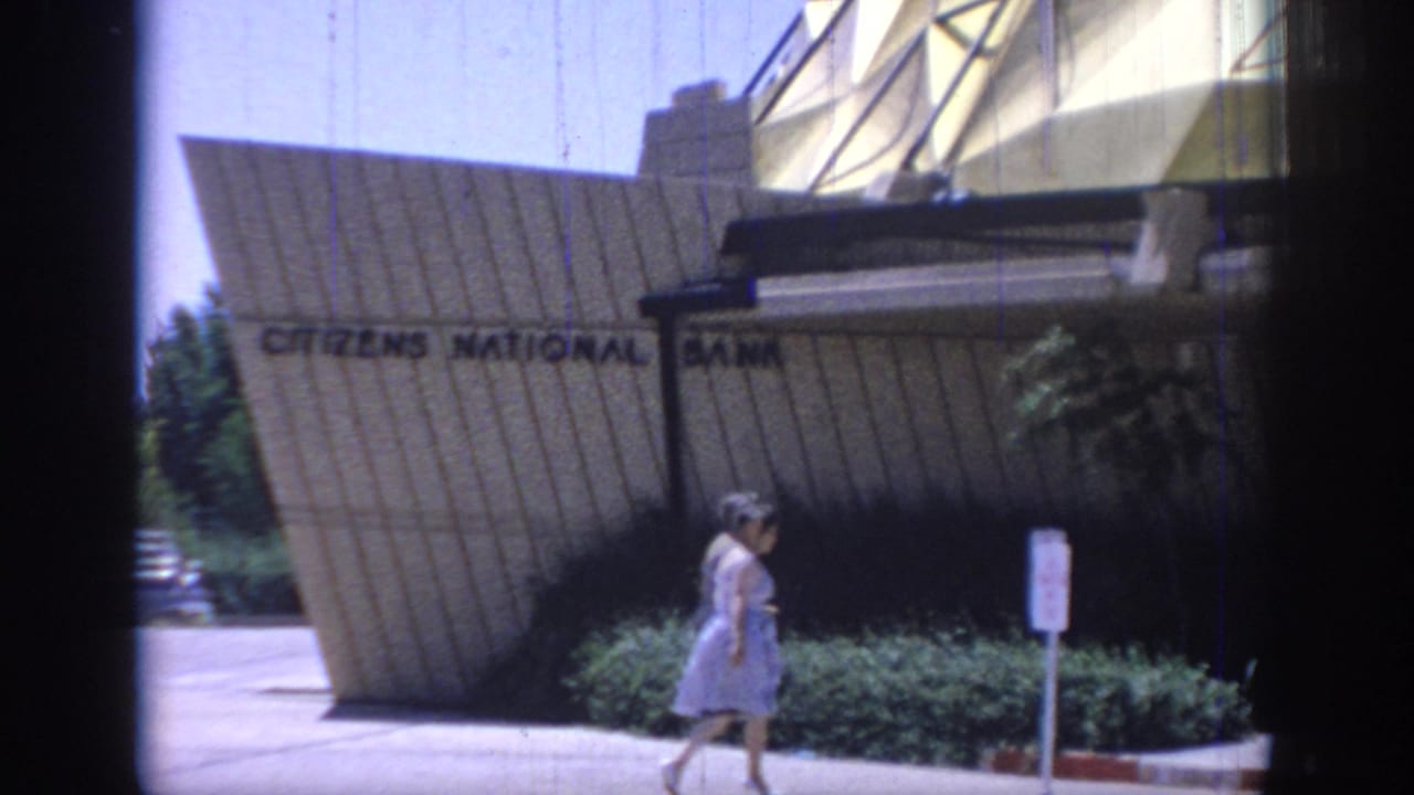 Oklahoma 1967 home-movie frame showing Citizens National Bank signage below the dome, with shrubs and a woman passing in front.