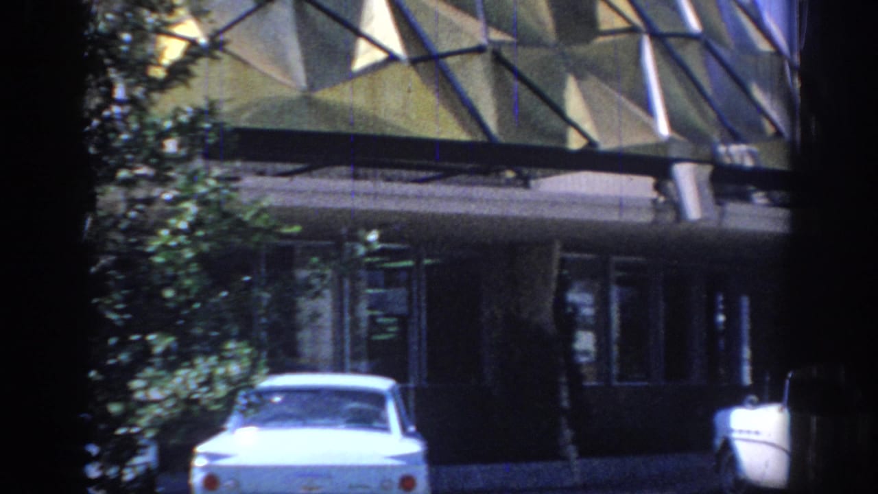 1967 Oklahoma 8mm frame showing a white 1960s car, leafy trees, and a modernist glass-fronted building exterior.