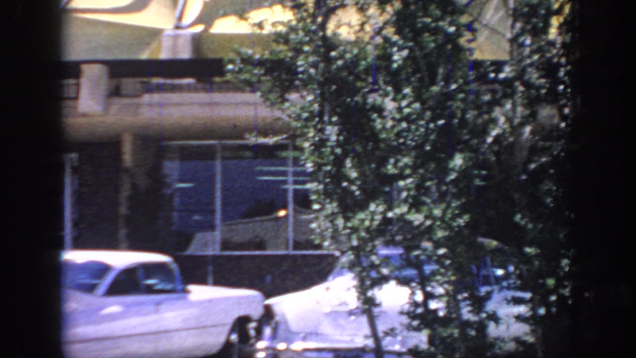 1967 Oklahoma 8mm frame with parked 1960s cars, tree cover, windows, and the lower facade of a modern building.