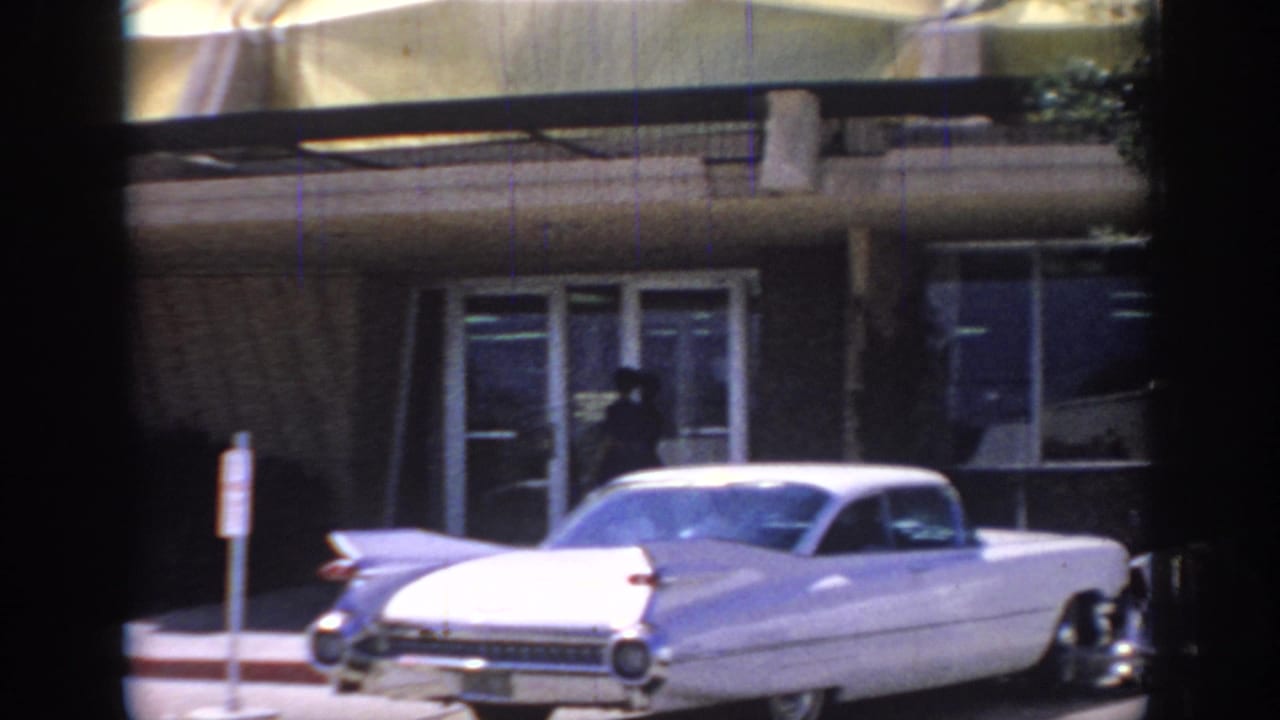 1967 Oklahoma street frame showing a large white 1960s sedan, storefront-style windows, and a modernist building exterior.
