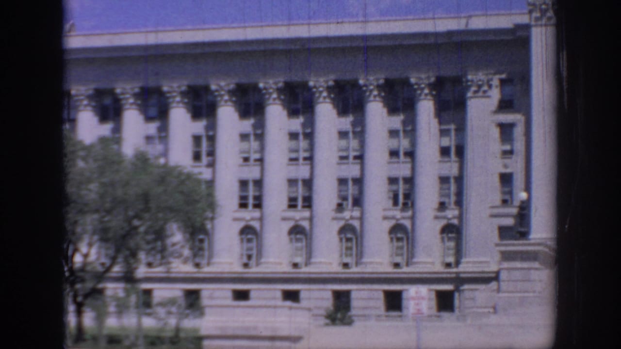 1967 Oklahoma State Capitol facade seen in 8mm footage, with white columns, rows of windows, trees, and blue sky.