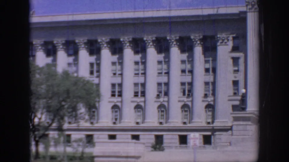 1967 Oklahoma State Capitol facade seen in 8mm footage, with white columns, rows of windows, trees, and blue sky.