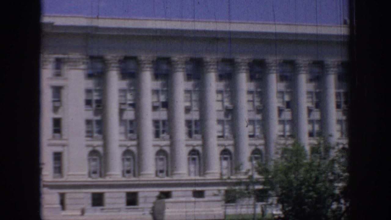 1967 Oklahoma 8mm view of a large classical facade with repeated columns, arched windows, blue sky, and tree foliage at the edge.