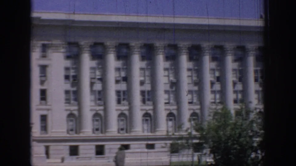 1967 Oklahoma 8mm view of a large classical facade with repeated columns, arched windows, blue sky, and tree foliage at the edge.