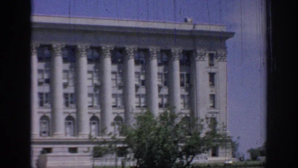 1967 Oklahoma 8mm frame with a columned public-looking building partly obscured by a leafy foreground tree and dark film borders.
