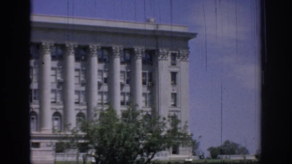 1967 Oklahoma 8mm frame showing the right side of a grand columned facade, foreground tree branches, and pale blue sky.