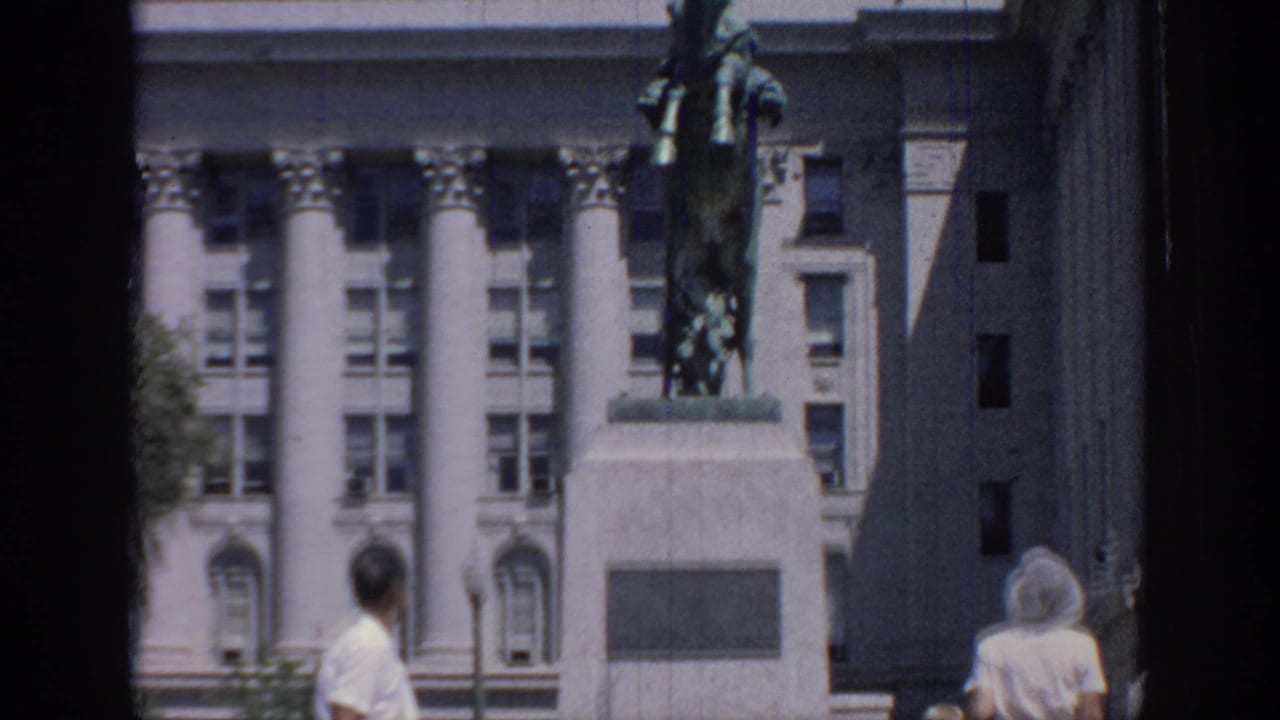 1967 Oklahoma home-movie frame showing a rearing horse statue before a classical columned building as visitors pass in the foreground.