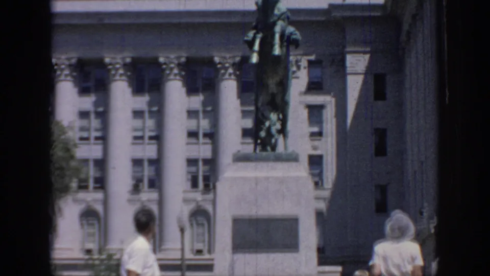 1967 Oklahoma home-movie frame showing a rearing horse statue before a classical columned building as visitors pass in the foreground.