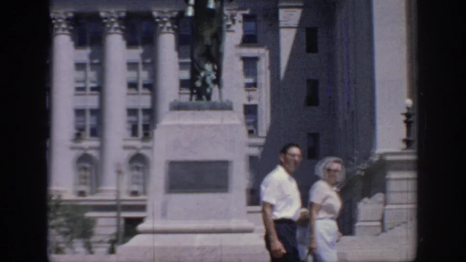 Two adults walk past the statue pedestal in a 1967 Oklahoma home-movie frame, with columns and city buildings behind them.