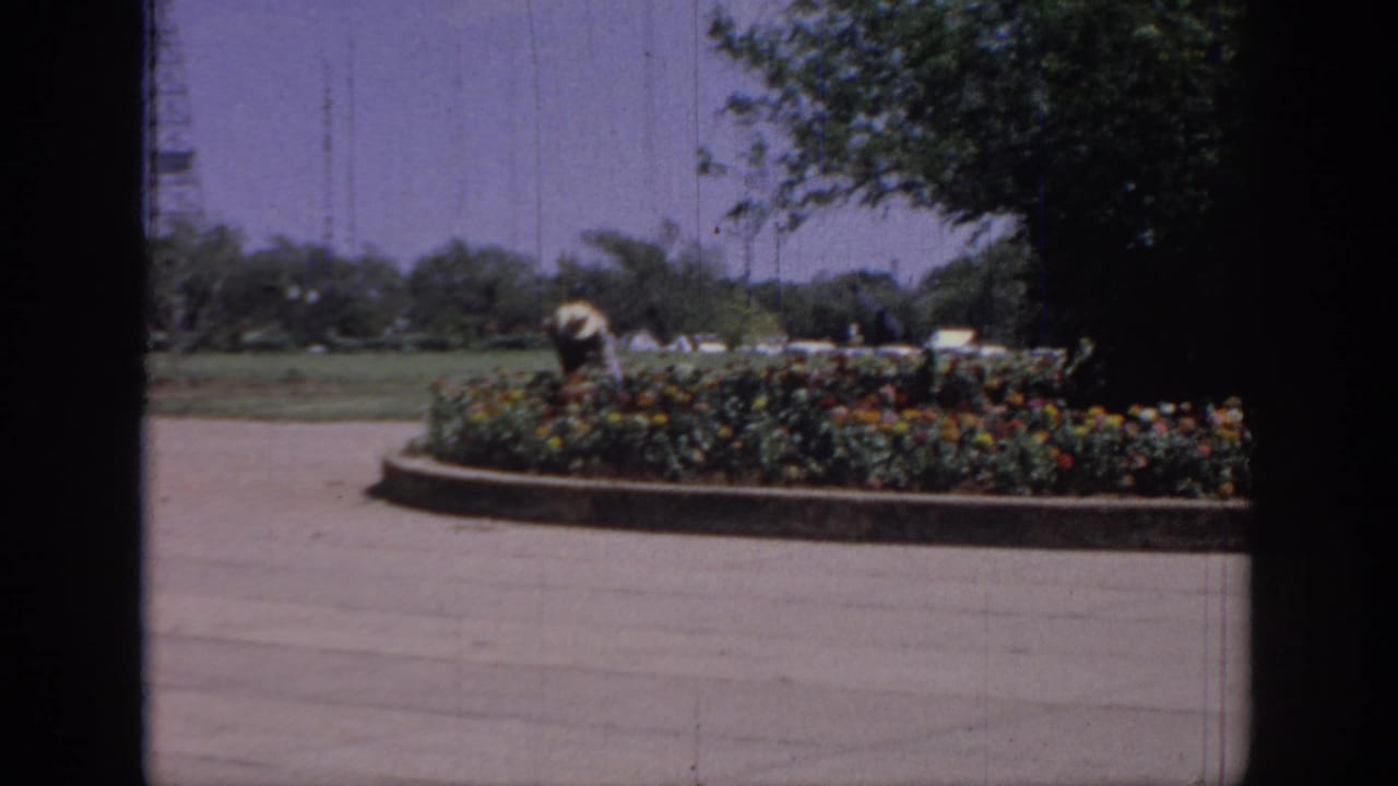 1967 Oklahoma 8mm frame showing a distant person in a light hat working near a circular flower bed, with trees and a utility tower behind.
